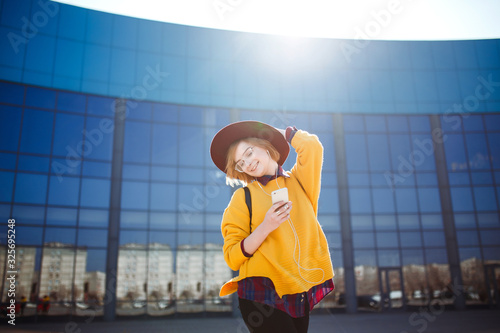 Young teenage girl with phone in her hands posing on buildings background. Hipster girl in a yellow sweater, hat and sunglasses using phone on a city street. Lifestyle, youth concept.