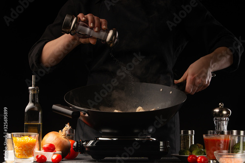 The chef cooks. Fries the dish in a frying pan. Salts the dish. frozen in motion. Restaurant business, on a black background for design, horizontal photo. Ingredients, cafe or restaurant business