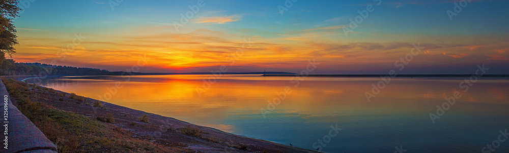 Fototapeta premium Panorama of colorful blue and golden sunset over the Kremenchug water reservoir in Svetlovodsk city, Kirovograd region, Ukraine. Beautiful romantic sunset view of the sea with sun reflection on water