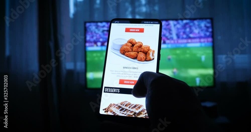 Background of the TV at home (at the bar), the guy orders different snacks, pizza and sweets scrolling through internet stores using mobile phones. Concept: Sport.