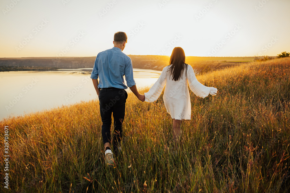 Happy fashionable couple in love holding hands and walking on coast near river. Girl clothes white dress. In background is sunset.