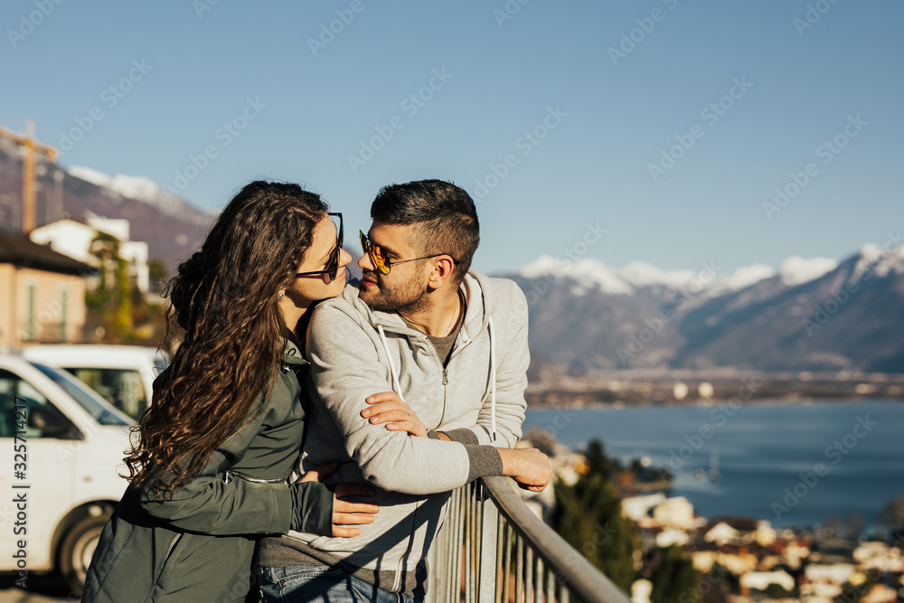 Beautiful couple in stylish clothes with glasses walking on snowy mountain background. Lovely couple travelling together in the Switzerland and walking around the lake Lugano.