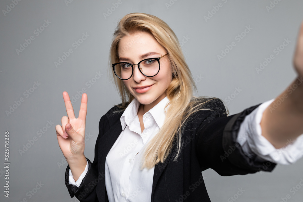 Portrait of business woman making selfie using smart phone and internet, gesturing peace symbol, having video call online over grey background Portrait of business woman making selfie using smart phone and internet, gesturing peace symbol, having video call online over grey background
