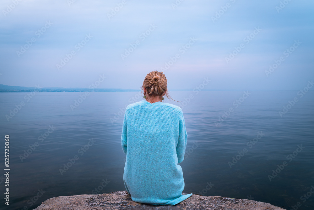 Beautiful single solo young traveler blond woman with tranquit jacket sitting alone on stone looking in to water in the blue ocean foggy mist lake in cold winter with sad dark depression  emotional