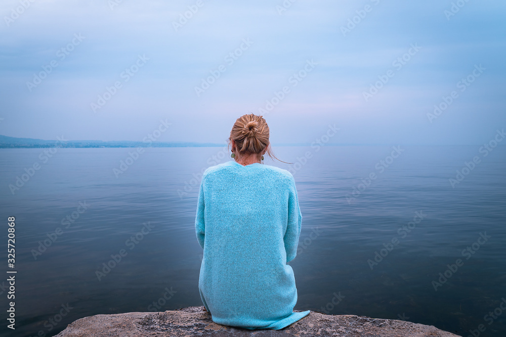 Fototapeta premium Beautiful single solo young traveler blond woman with tranquit jacket sitting alone on stone looking in to water in the blue ocean foggy mist lake in cold winter with sad dark depression emotional