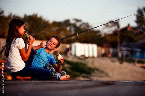 Two young cute little friends, boy and girl eating sandwiches and fishing, boy shoving gesture thumb up