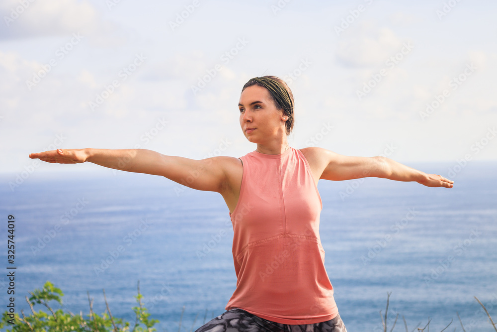 Close up warrior pose. Young woman practicing yoga, standing in Virabhadrasana pose. Outdoor yoga on the cliff. Bali, Indonesia Close up warrior pose. Young woman practicing yoga, standing in Virabhadrasana pose. Outdoor yoga on the cliff. Bali, Indonesia