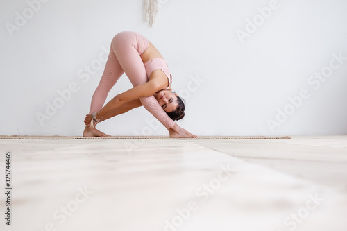 Pacified calm young woman in a pink suit doing parshvottanasana while standing barefoot on a rug on the floor against a white background with wicker macram? on the wall. Place for advertising photo