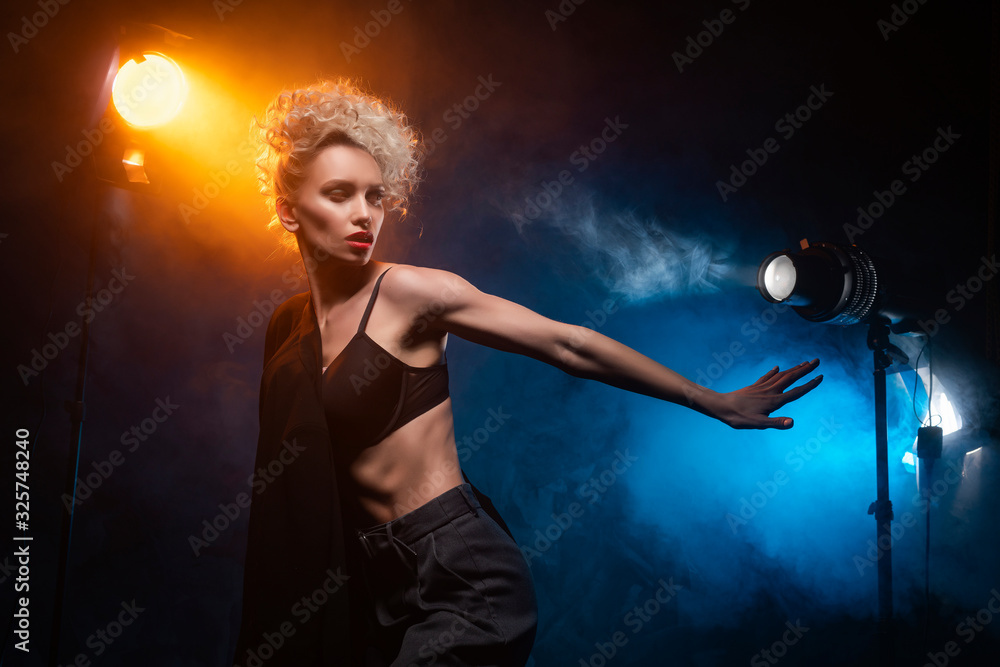 A beautiful blonde girl with an elegant hairstyle and large breasts, wearing a bra, trousers and a blazer, artistically poses in the rays of spotlights in the smoke. Cinematic, art, commercial design.
