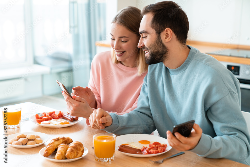 Portrait of young joyful couple using cellphones while having breakfast
