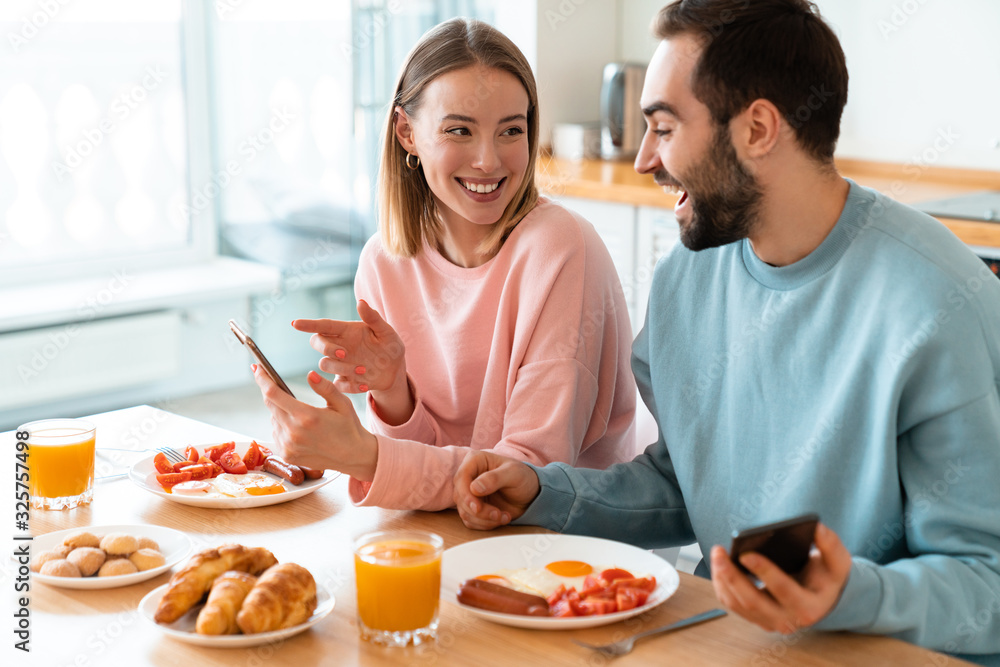 Portrait of young joyful couple using cellphones while having breakfast