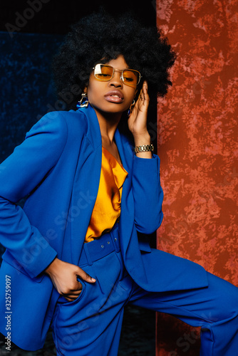 African American fashionable woman wearing yellow sunglasses, top, classic blue suit with blazer, high waist trousers. Young beautiful confident model posing near colorful walls. Fashion portrait photo