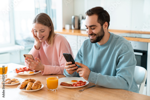 Portrait of young joyful couple using cellphones while having breakfast