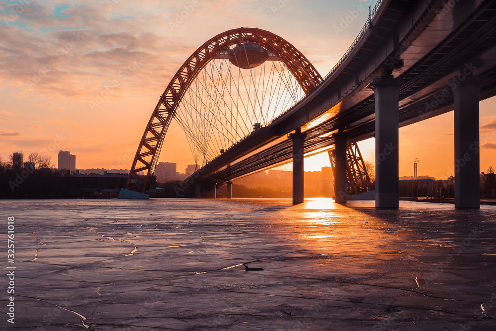 Obraz premium The photo shows picturesque bridge with big red arch over the river. This cable-stayed bridge stands on the frozen Moscow river. Crimson sunny rays illuminate bridge details and ice on the sunset. 