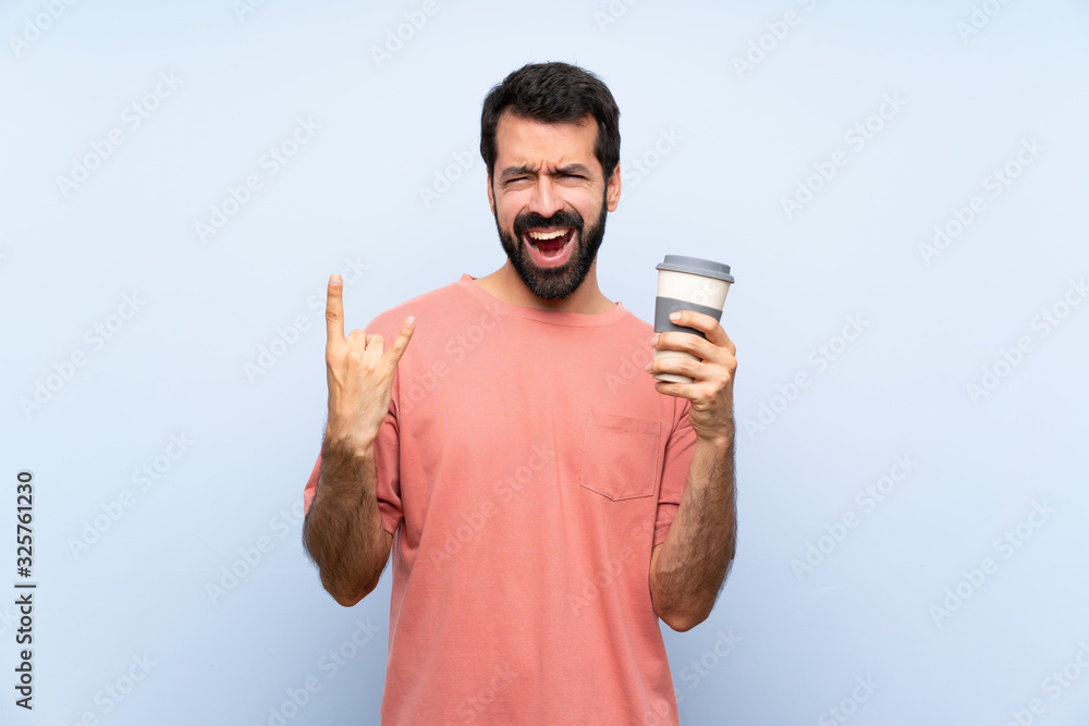Young man with beard holding a take away coffee over isolated blue background making rock gesture Young man with beard holding a take away coffee over isolated blue background making rock gesture