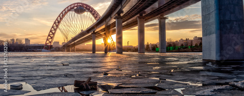 Panoramic HDR photo shows picturesque bridge with big red arch over the river. This cable-stayed bridge stands on the frozen Moscow river. Sunny rays illuminate bridge details and ice on the sunset. photo