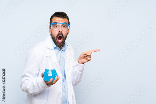 Young scientific holding laboratory flask over isolated background surprised and pointing side