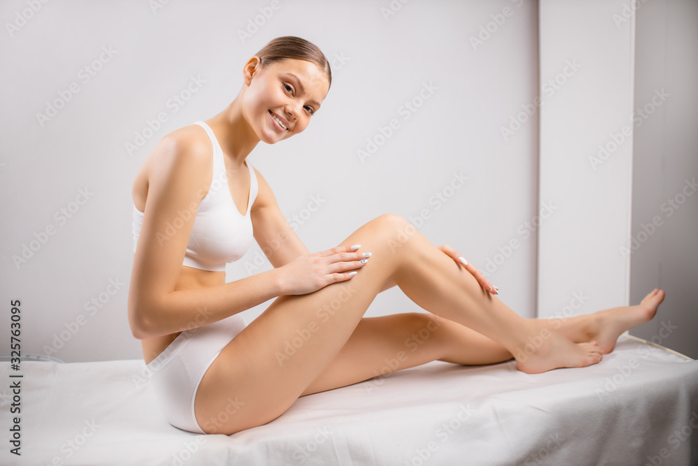 portrait of beautiful young caucasian woman enjoy her soft, smooth and hairless skin after epilation. woman after hair removal from legs, sit on bed in underwear and look at camera portrait of beautiful young caucasian woman enjoy her soft, smooth and hairless skin after epilation. woman after hair removal from legs, sit on bed in underwear and look at camera