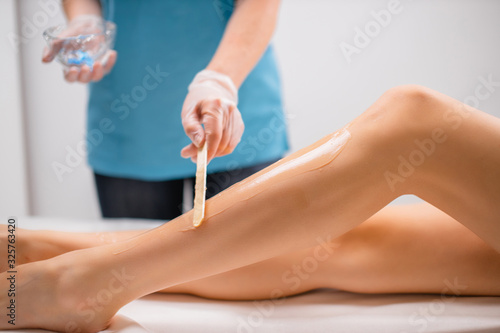 side view on professional beautician applying moisturizer on female legs before laser epilation procedure. slender woman lie on bed, she want soft, hairless and smooth skin