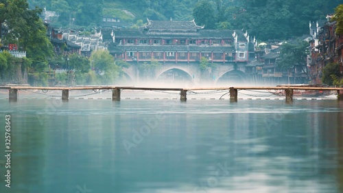 A simple wooden bridge on a river canal with buildings in the background. Fenghuang County, Hunan, China