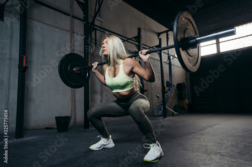 Young blonde woman with barbell flexing muscles and making shoulder press squat.