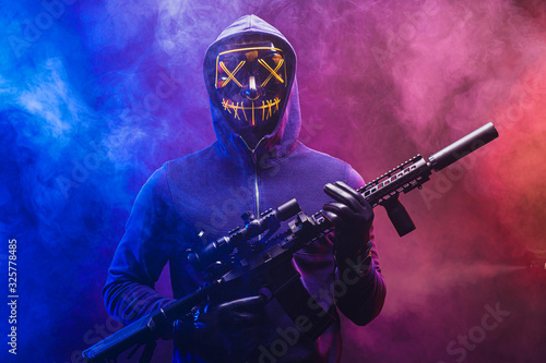 confident dangerous killer man in pullover stand in the hood with weapon, masked hunter man isolated over smoky space with neon lights. crime, kill, hunt, danger concept