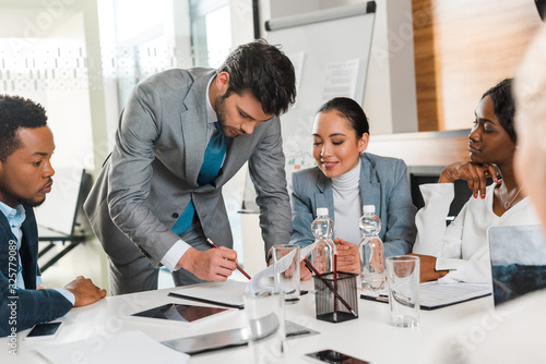 selective focus of handsome businessman holding pencil while standing near multicultural colleagues sitting at desk in conference hall