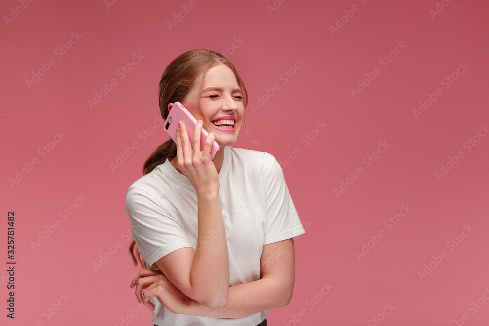 Portrait of cute happy redhead girl wearing white t-shirt talking on mobile phone and smiling isolated over pink background. Copy space. Young people working with mobile devices. Portrait of cute happy redhead girl wearing white t-shirt talking on mobile phone and smiling isolated over pink background. Copy space. Young people working with mobile devices.
