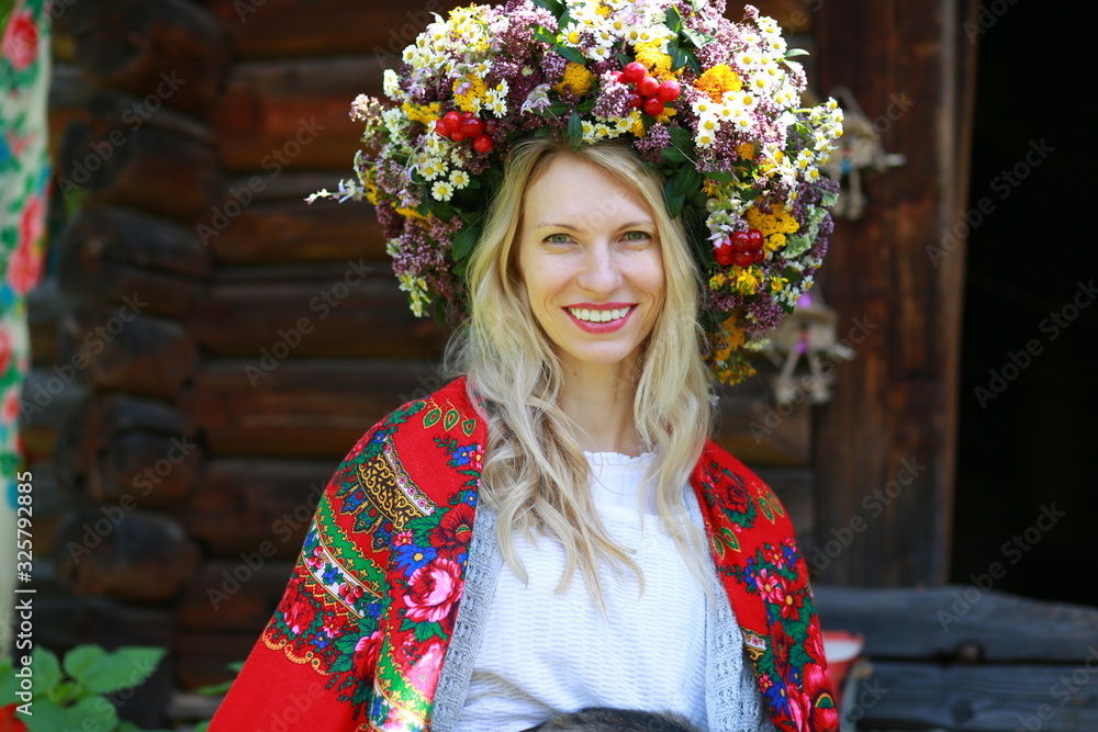 beautiful young girl sitting on the porch near a wooden house in a wreath of fresh flowers and a red scarf on her shoulders, Ukrainian national ethnic style