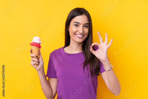 Young brunette woman with a cornet ice cream over isolated yellow background showing ok sign with fingers © luismolinero