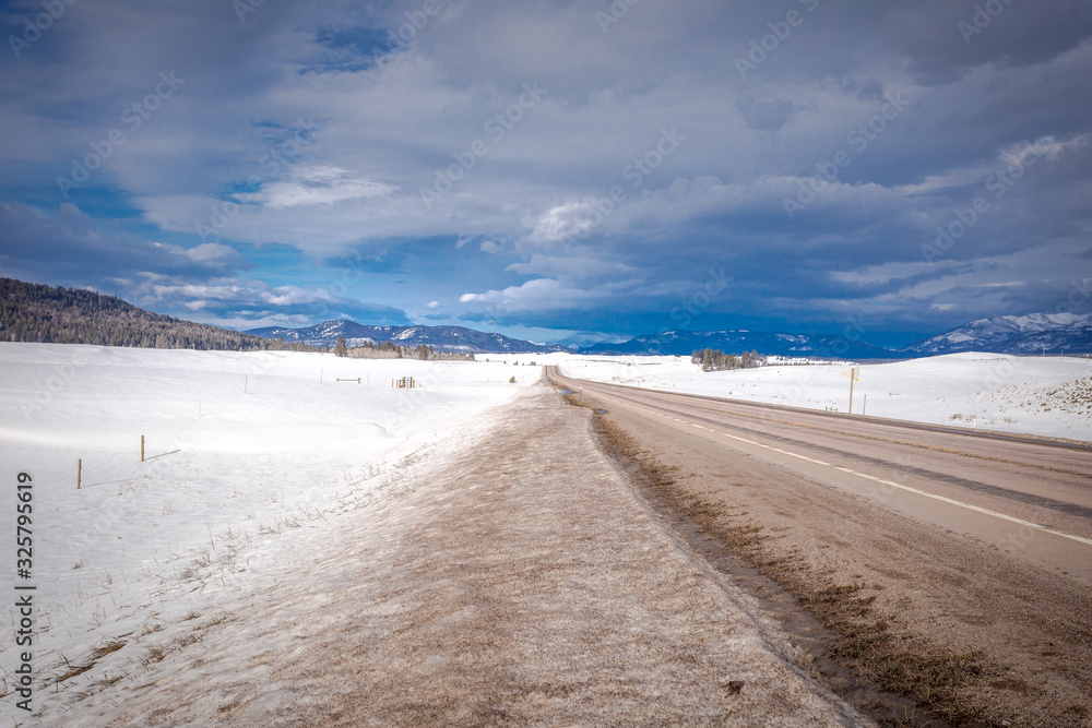 Empty highway in wide open area of Montana covered in snow with mountain range on cloudy day