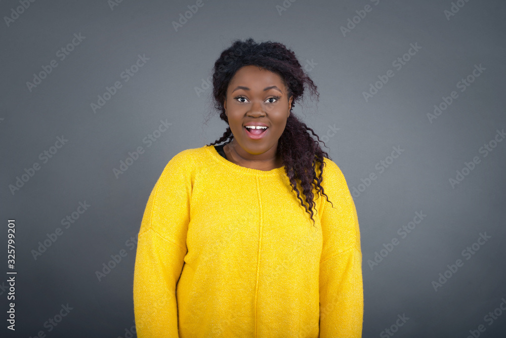 Emotive young beautiful woman wears casual clothes, happy and excited to celebrate anniversary. Happy African American woman has good mood stands against gray wall. Emotive young beautiful woman wears casual clothes, happy and excited to celebrate anniversary. Happy African American woman has good mood stands against gray wall.