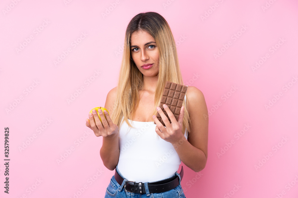 Young Uruguayan woman over isolated pink background taking a chocolate tablet in one hand and an apple in the other Young Uruguayan woman over isolated pink background taking a chocolate tablet in one hand and an apple in the other