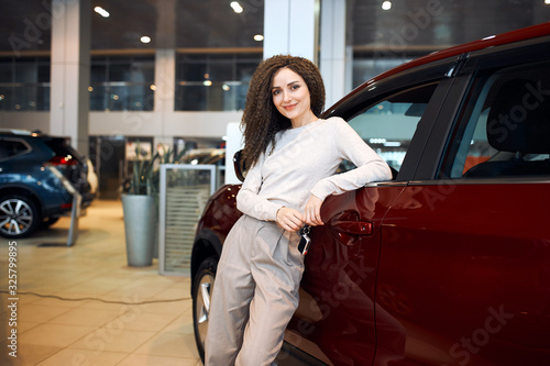 Beautiful young business woman in stylish clothes is smiling and looking at camera while leaning on a red car in a motor show, close up photo, sale , discount, trade, industry © the faces