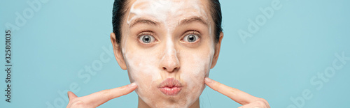 panoramic shot of funny woman with cleansing foam on face pointing on cheeks, isolated on blue