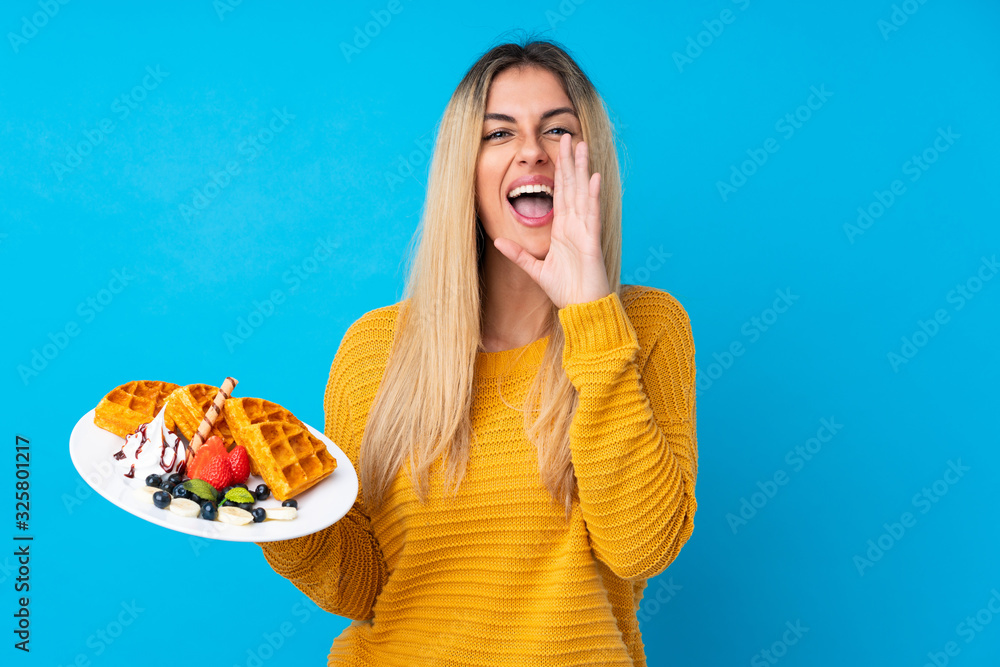 Young woman holding waffles over isolated background shouting with mouth wide open Young woman holding waffles over isolated background shouting with mouth wide open