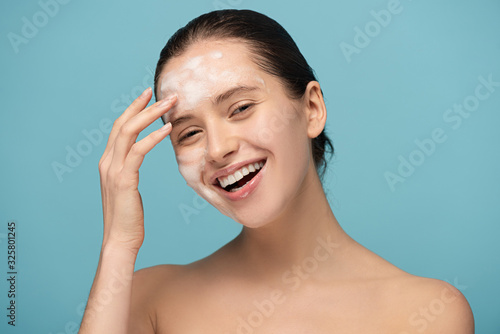 laughing young woman applying cleansing foam on face, isolated on blue