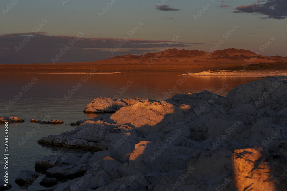Large sharp stones lie in the lake and are illuminated by the orange light of the setting sun. Bright blue stars on a blue sky with clouds. Traveled photo in Mongolia. 