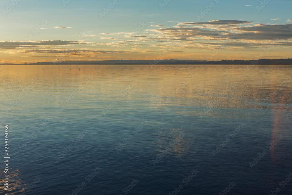 The smooth calm surface of the lake is lit by the light of the setting sun. The sky in orange tones is reflected in the water of the lake. Traveled photo in Mongolia.
