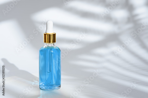trendy dropper glass bottle with pipette and blue liquid on white background with leaf shadow