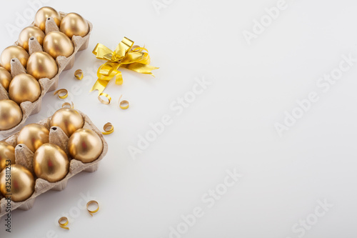 A dozen golden eggs decorated with a gold bow, on the edge of a white background with place for text. Concept backgrounds for Easter.