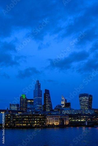 City of London in the UK. View over the River Thames to the city of London in the evening. Cityscape of the CBD with skyscrapers, lights, river and blue sky with clouds at night. London, England 2020.