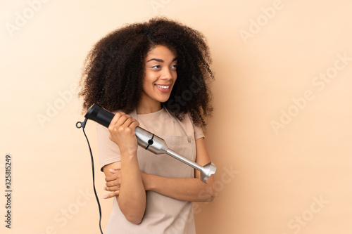 Young african american woman using hand blender isolated on beige background looking to the side