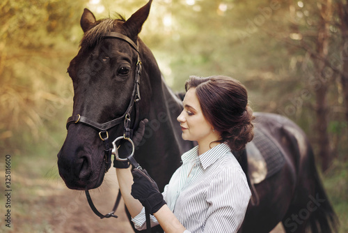 Beautiful girl jockey rider with a black horse, dressed in a light shirt in a green forest Park. The concept of horse riding.