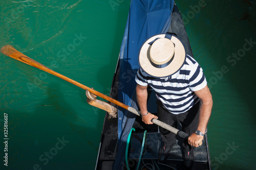 Overhead view of Venetian gondolier in traditional hat and striped shirt rowing a gondola under a bridge on a canal in Venice, Italy