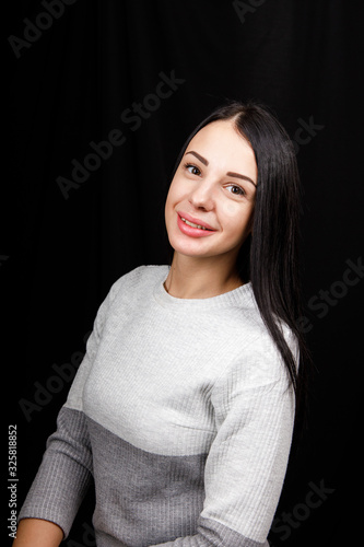 Portrait of serious beautiful female with black hair, has minimal makeup, looks calmly at camera, wears white jumper, stands against black background, being deep in thoughts