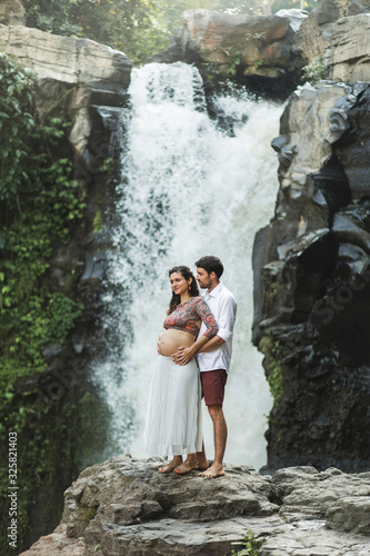 Childbirth in Bali. Young pregnant couple travel on Tegenungan waterfall. Family harmony values. Happy together, pregnancy travel lifestyle. Beautiful morning nature. Maternity concept.