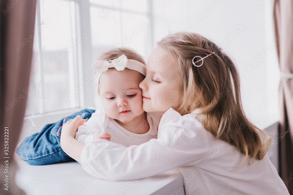 Two cute sisters sitting near window close to each other and playing. Two cute sisters sitting near window close to each other and playing.
