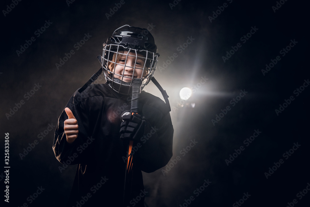 Young blonde sporty boy, ice hockey player, posing in a dark studio for a photoshoot, wearing an ice-skating uniform, helmet, hockey stick, showing a good gesture with his hand and smiling on camera