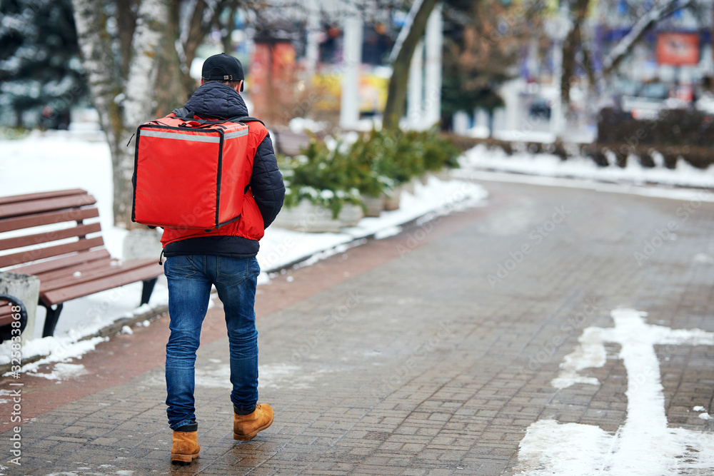 Courier is delivering food with red thermal backpack. Delivery boy goes and carries isothermal bag with ordered food. Fast delivery service from favorite restaurants in any weather around the clock